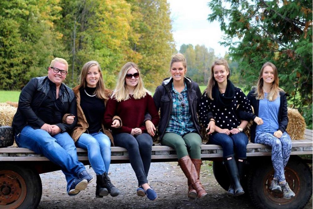 A group of 6 smiling people sitting on a flat trailer behind a truck, with fall foliage in the background