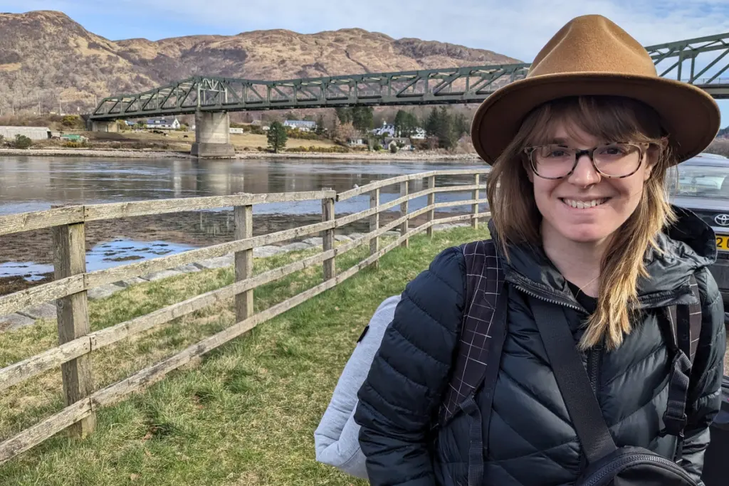 Taylor Bonin standing beside a river with a bridge and mountain landscape in the background.