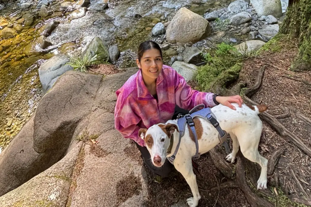 Ximena Vidaurre Leyva sitting beside a forest stream with her dog during an outdoor nature adventure.