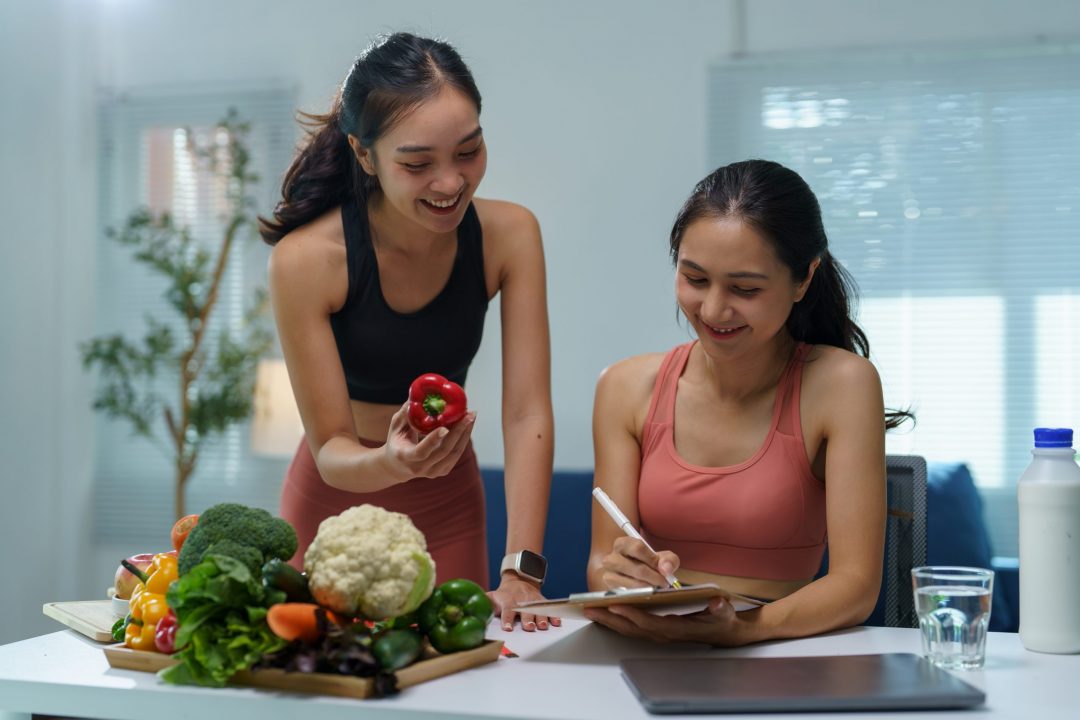 Two young women discussing a diet plan in a kitchen, with a nutritionist holding a red bell pepper and a client taking notes, surrounded by fresh vegetables